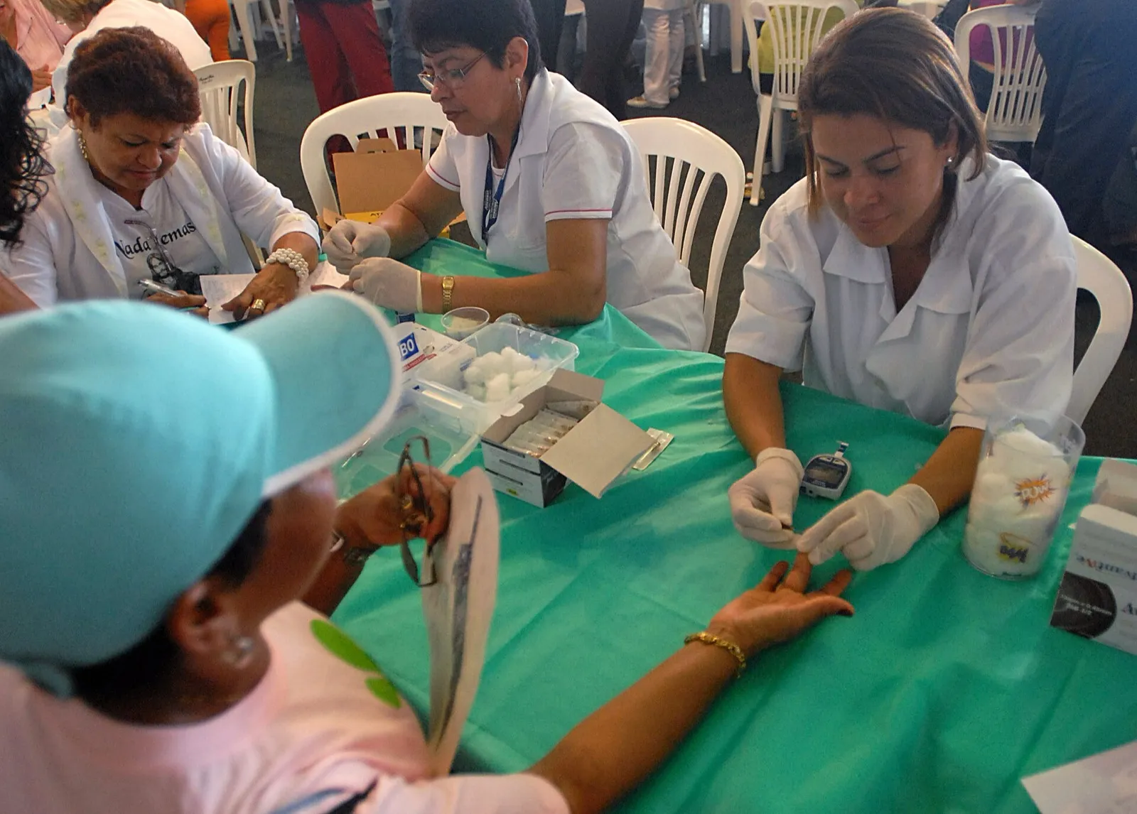 Profissional de saúde de luvas brancas faz teste de glicemia no dedo de uma participante, com um medidor sobre a mesa verde