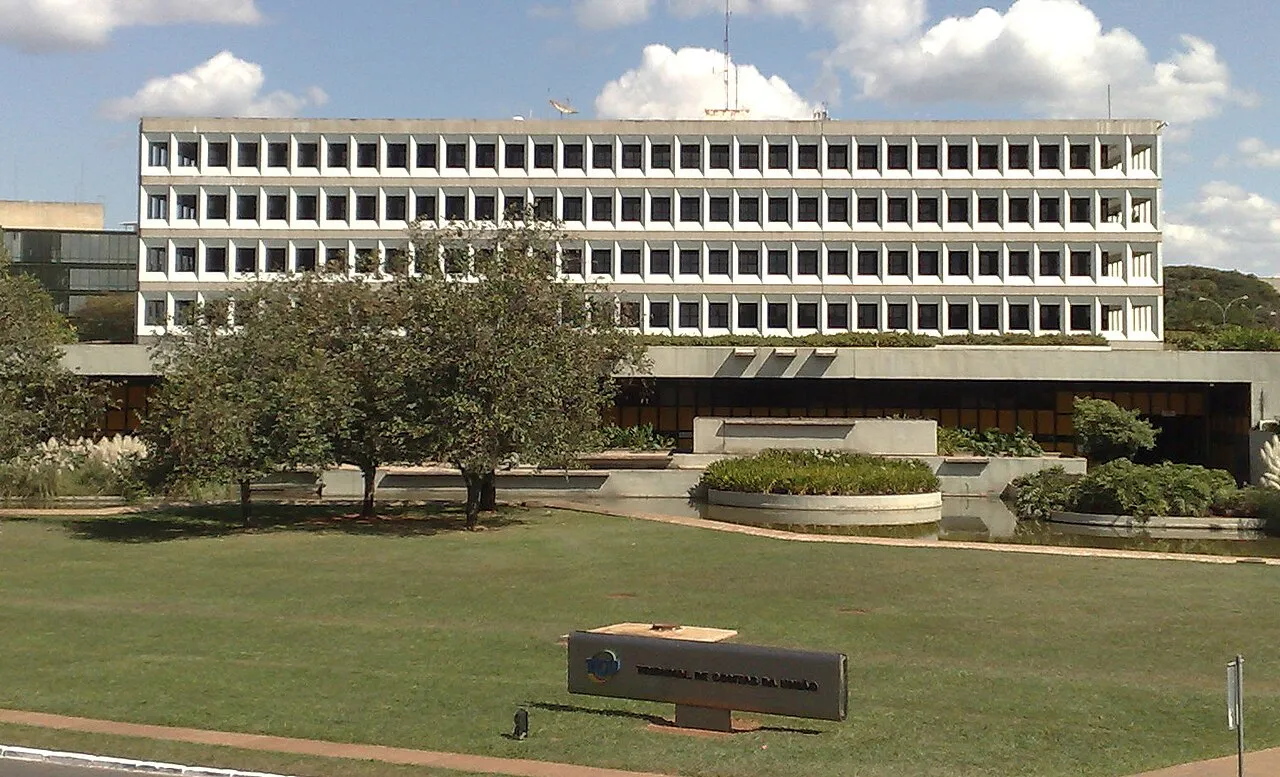 Prédio branco de cinco andares da sede do Tribunal de Contas da União, com gramado e uma placa com o nome do tribunal à frente.