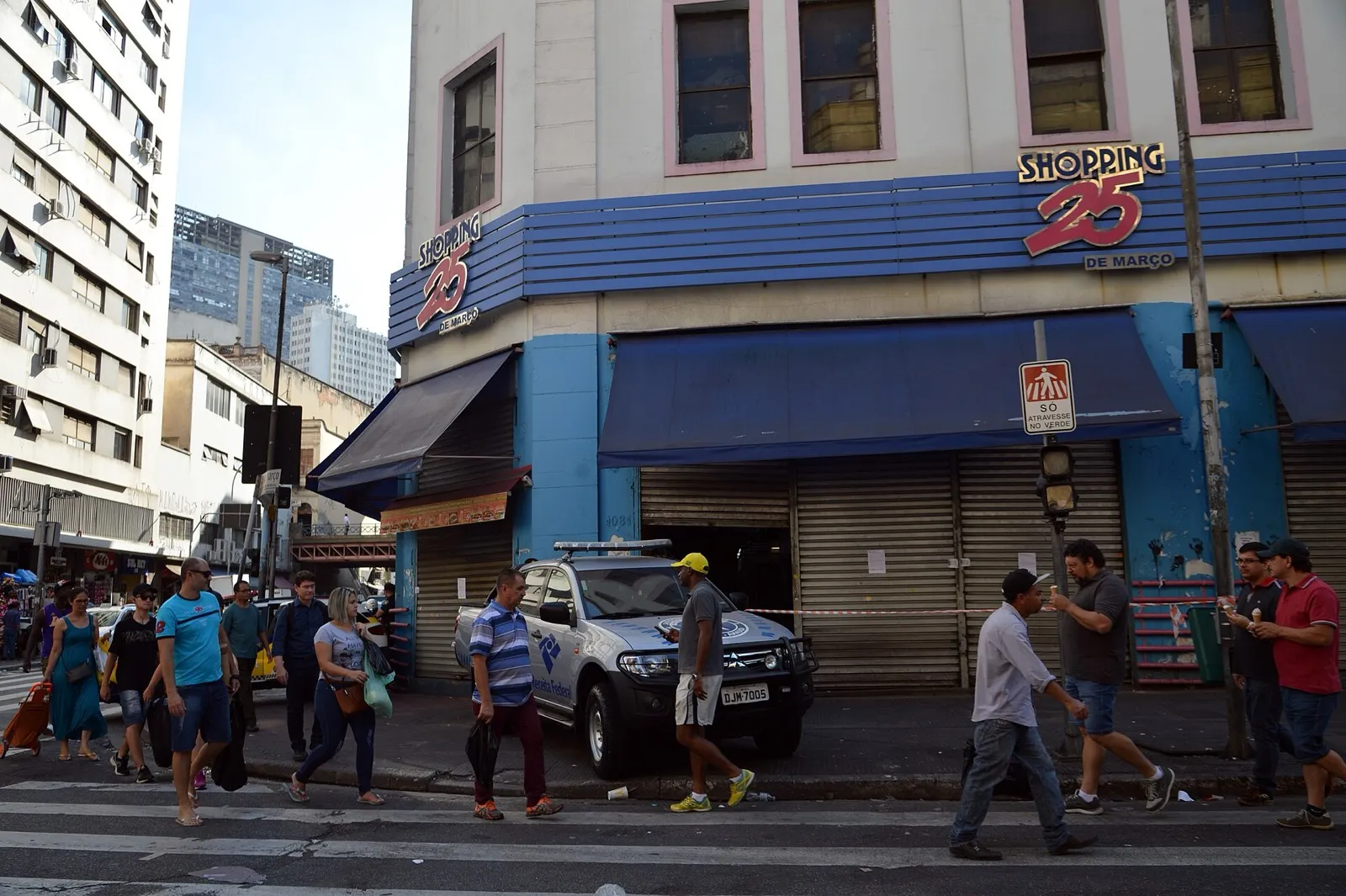 Fachada do Shopping 25 de Março, em São Paulo, com lojas de portas fechadas por grades, um carro da Receita Federal na calçada e pedestres atravessando a rua