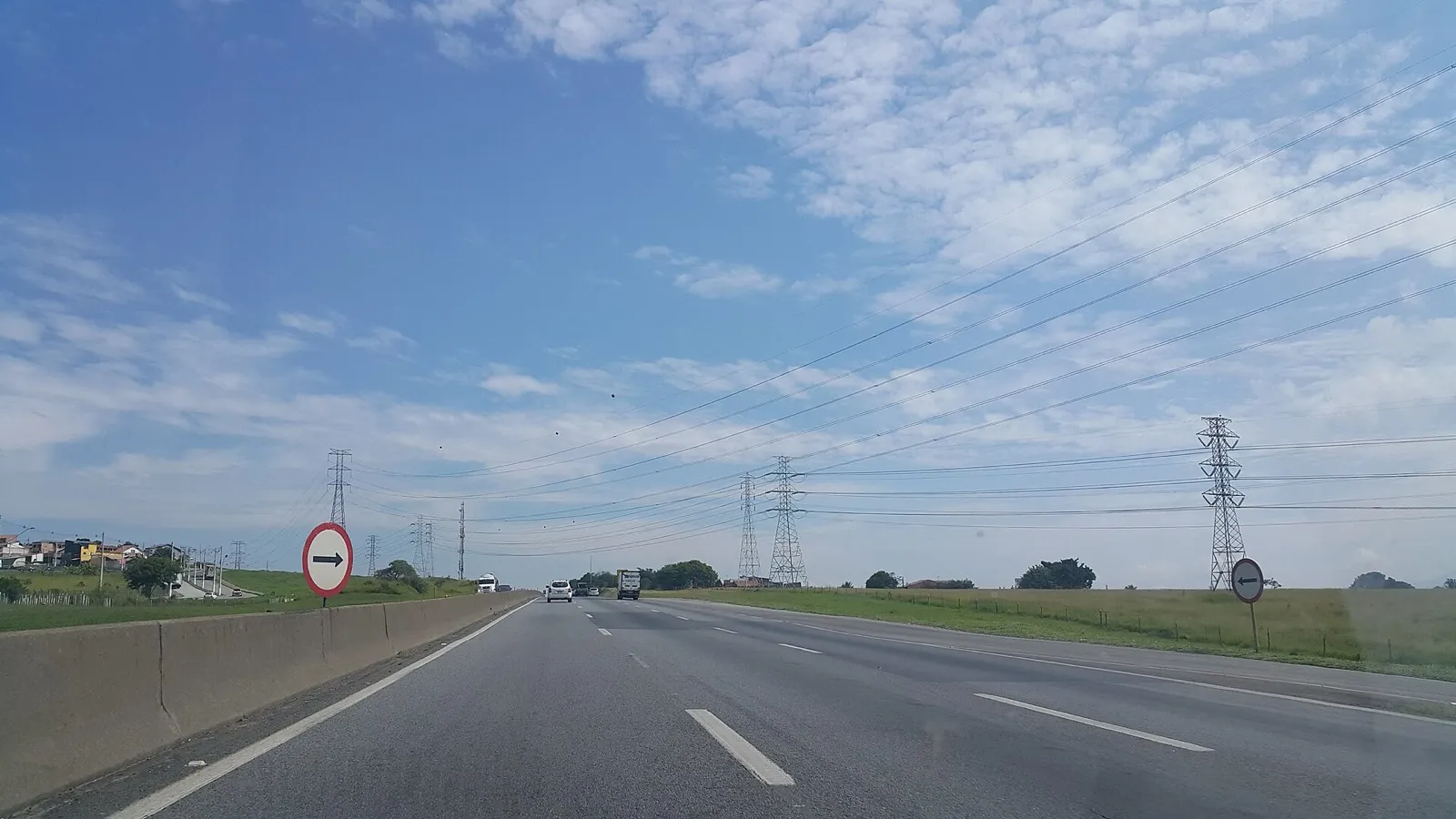 Pista de rodovia duplicada com poucos carros e um caminhão, mureta de concreto à esquerda, placas de sinalização e torres de transmissão de energia sob céu azul.