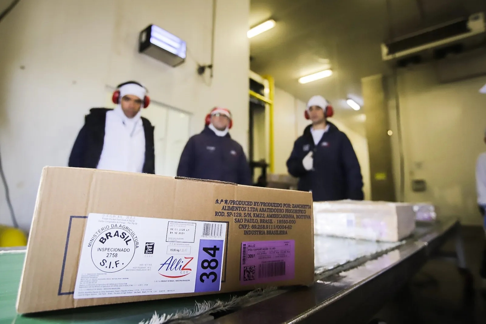 Caixa de frango congelado com selo de inspeção federal (S.I.F.) e rótulo em chinês, em primeiro plano, com três funcionários de touca e protetor auricular desfocados ao fundo, dentro de um frigorífico.