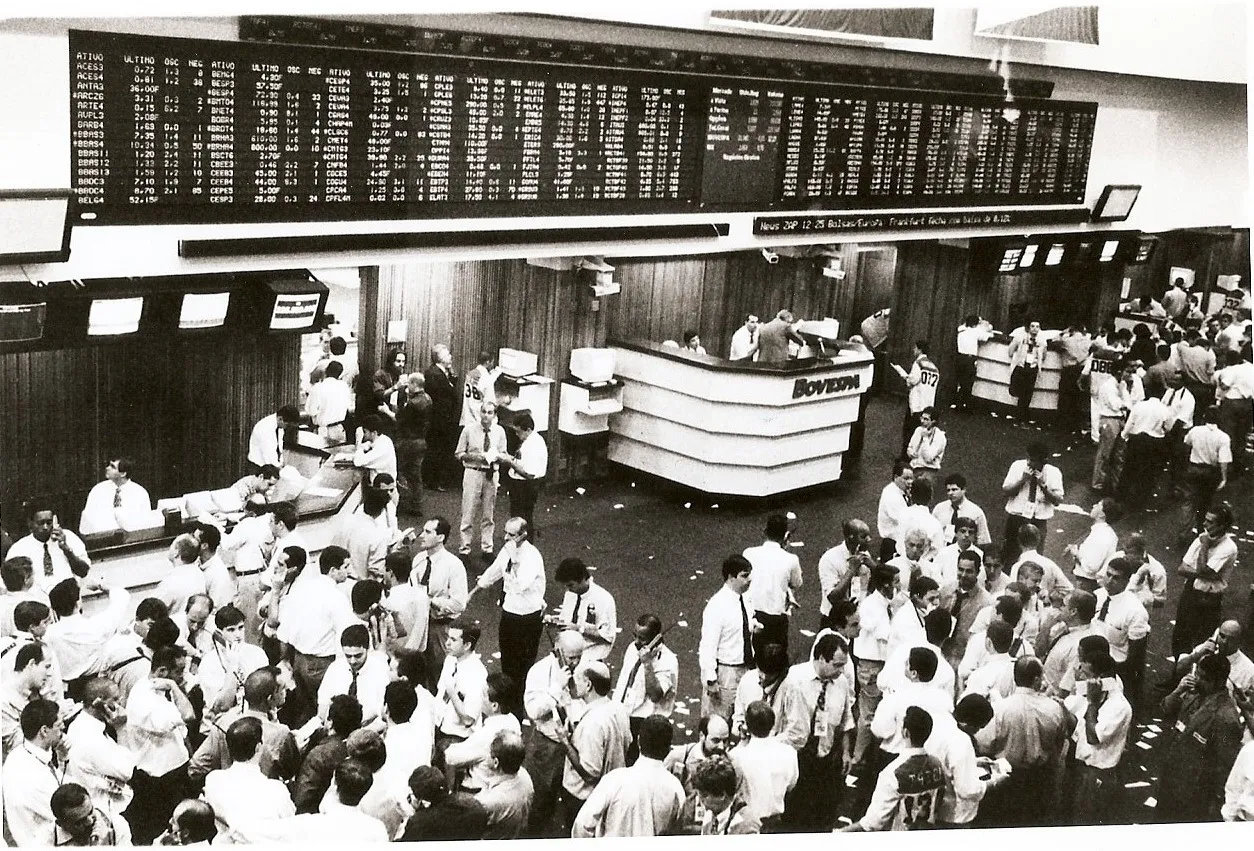 Foto em preto e branco do antigo pregão da Bovespa, com painel de cotações ao alto e dezenas de operadores no salão.