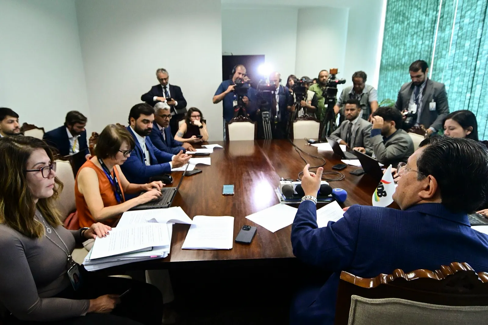Senador Eduardo Braga, de paletó azul, gesticula à mesa de reunião durante entrevista coletiva, com jornalistas e câmeras de TV ao fundo.