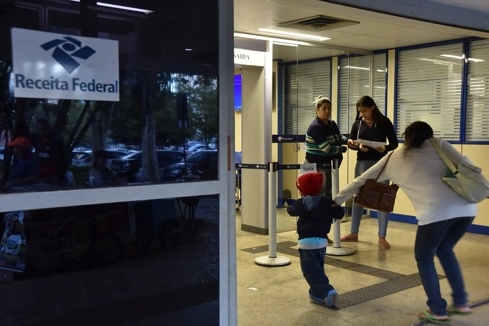 Pessoas na entrada de uma unidade da Receita Federal, com logotipo do órgão na porta de vidro