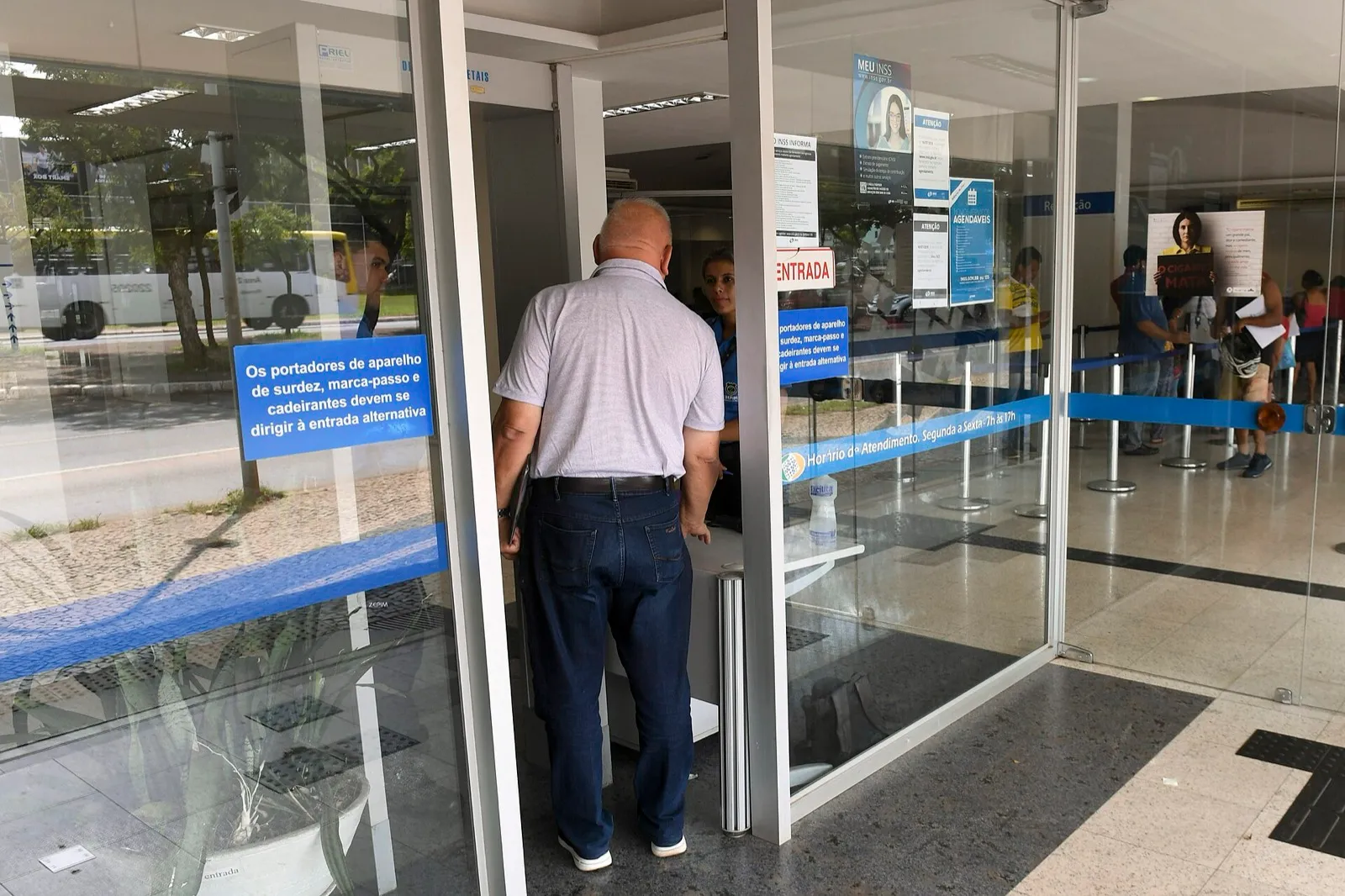 Entrada de uma agência do INSS: um homem de camisa clara entra pela porta de vidro enquanto uma atendente espera no balcão e pessoas aguardam em fila ao fundo