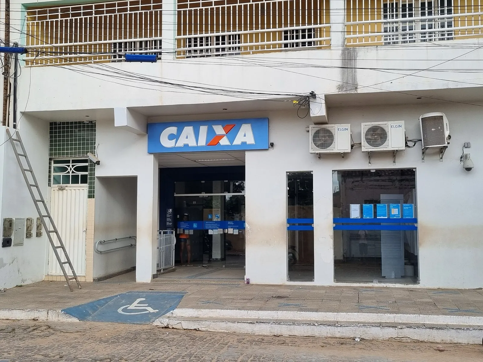 Fachada de uma agência da Caixa Econômica Federal com letreiro azul, porta de vidro e aparelhos de ar-condicionado na parede branca.