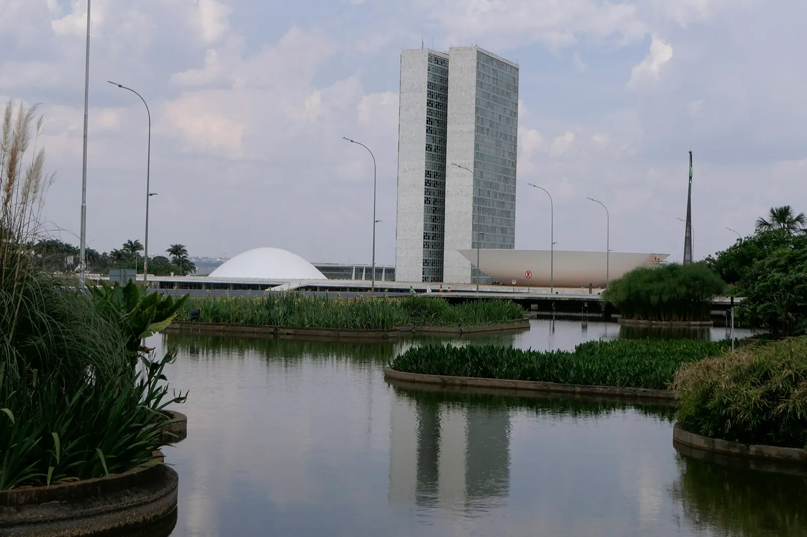 Sede do Congresso Nacional em Brasília, com as duas torres de gabinetes ao centro e as cúpulas do plenário ao fundo, refletidas em um espelho d'água cercado de vegetação.