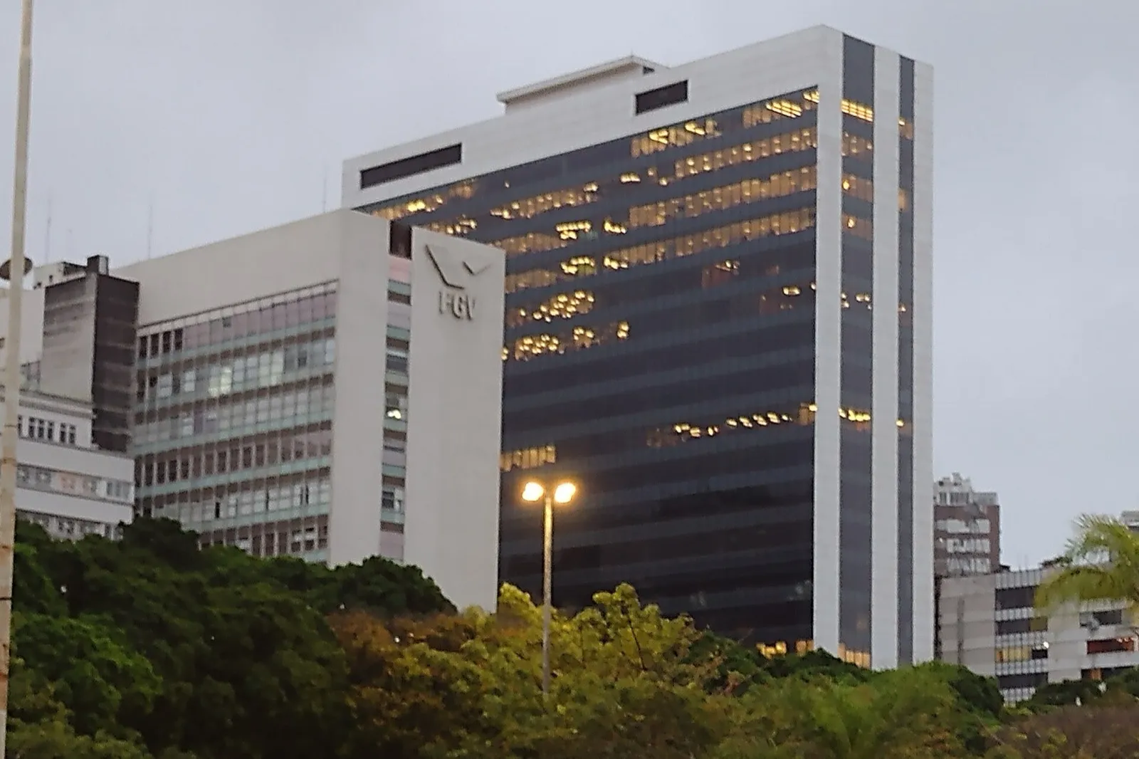 Fachada do prédio da Fundação Getulio Vargas no Rio de Janeiro, com a marca FGV em uma torre branca ao lado de uma torre espelhada, vista de uma avenida arborizada