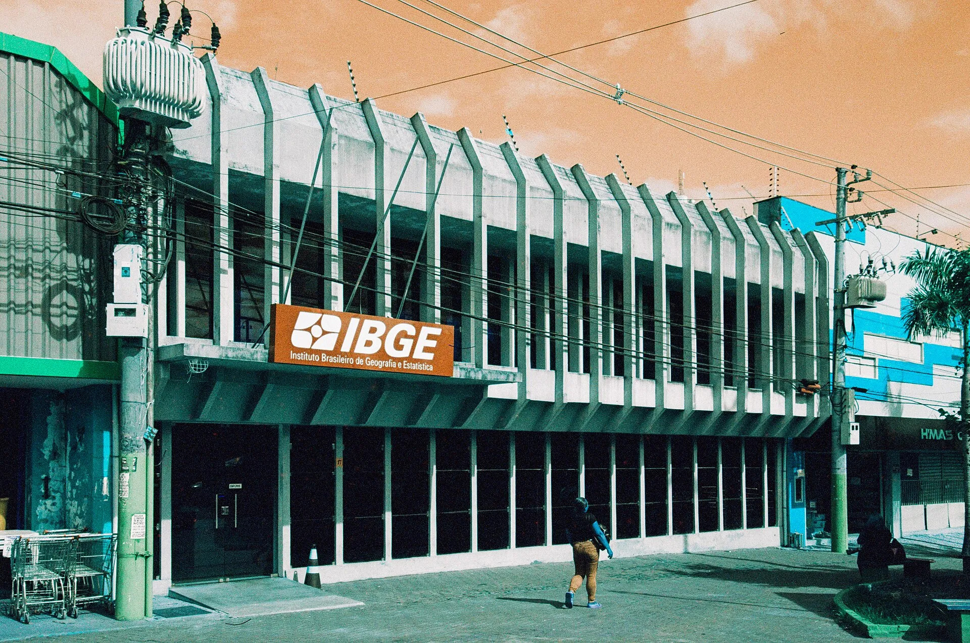 Fachada do prédio do IBGE em Cuiabá, com placa laranja e branca do instituto, em rua com fios elétricos e um pedestre.