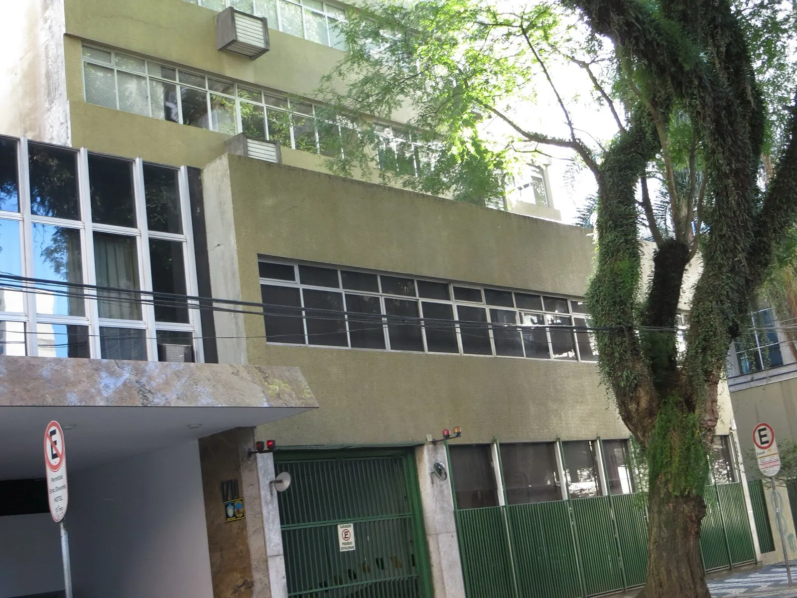 Fachada de prédio bege, com janelas horizontais, portão verde e uma árvore grande à direita, no Centro de Curitiba.