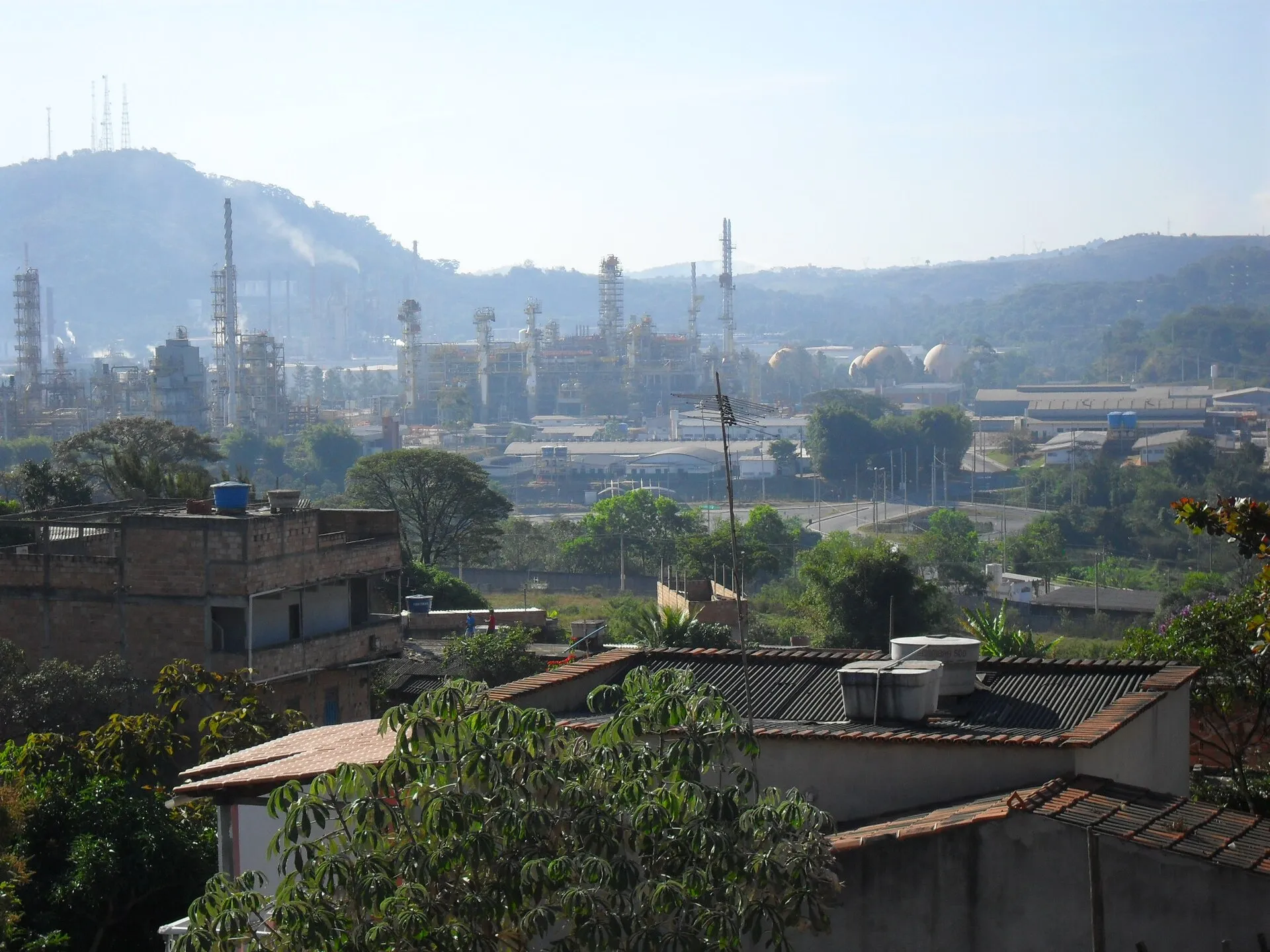 Vista de uma refinaria da Petrobras, com torres e colunas metálicas, cercada por casas, árvores e morros, em Ibirité (MG).