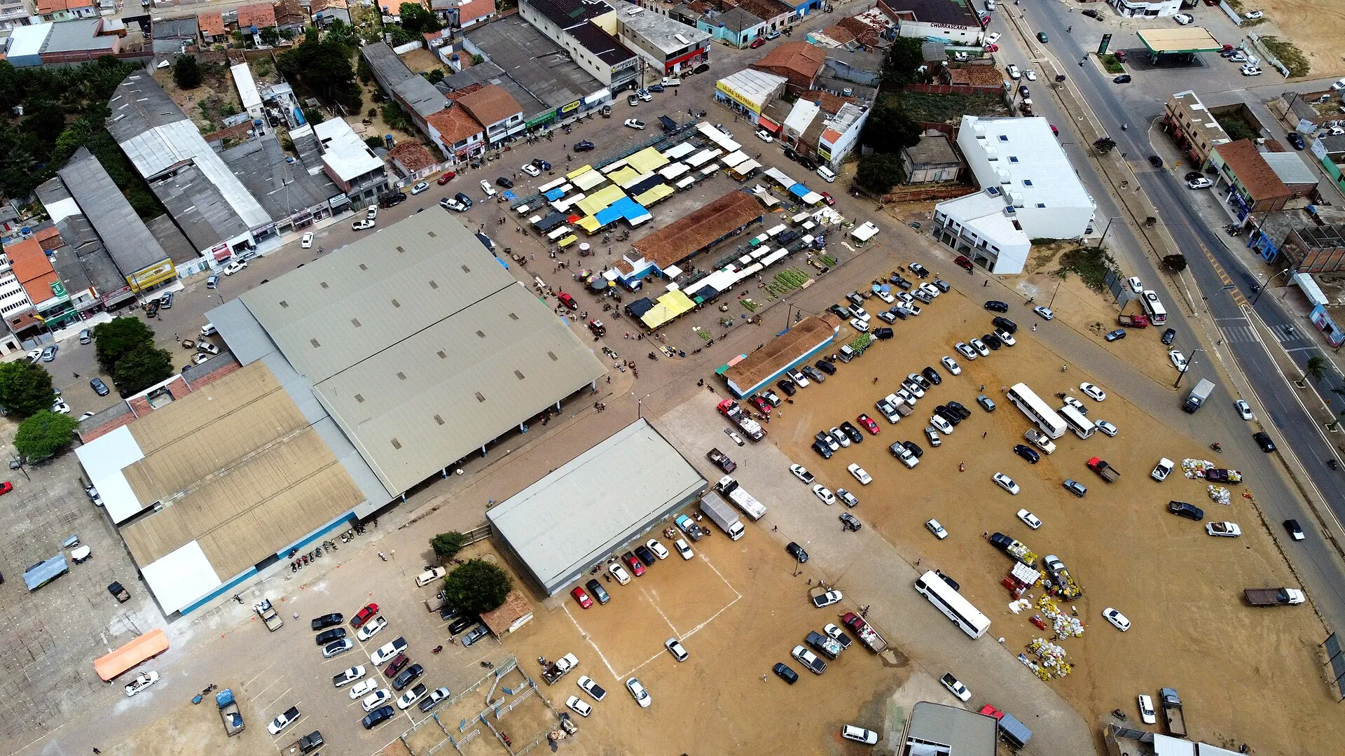 Vista aérea de feira livre em Maracás, na Bahia, com barracas coloridas, um galpão coberto, carros estacionados e ruas de uma cidade do interior.