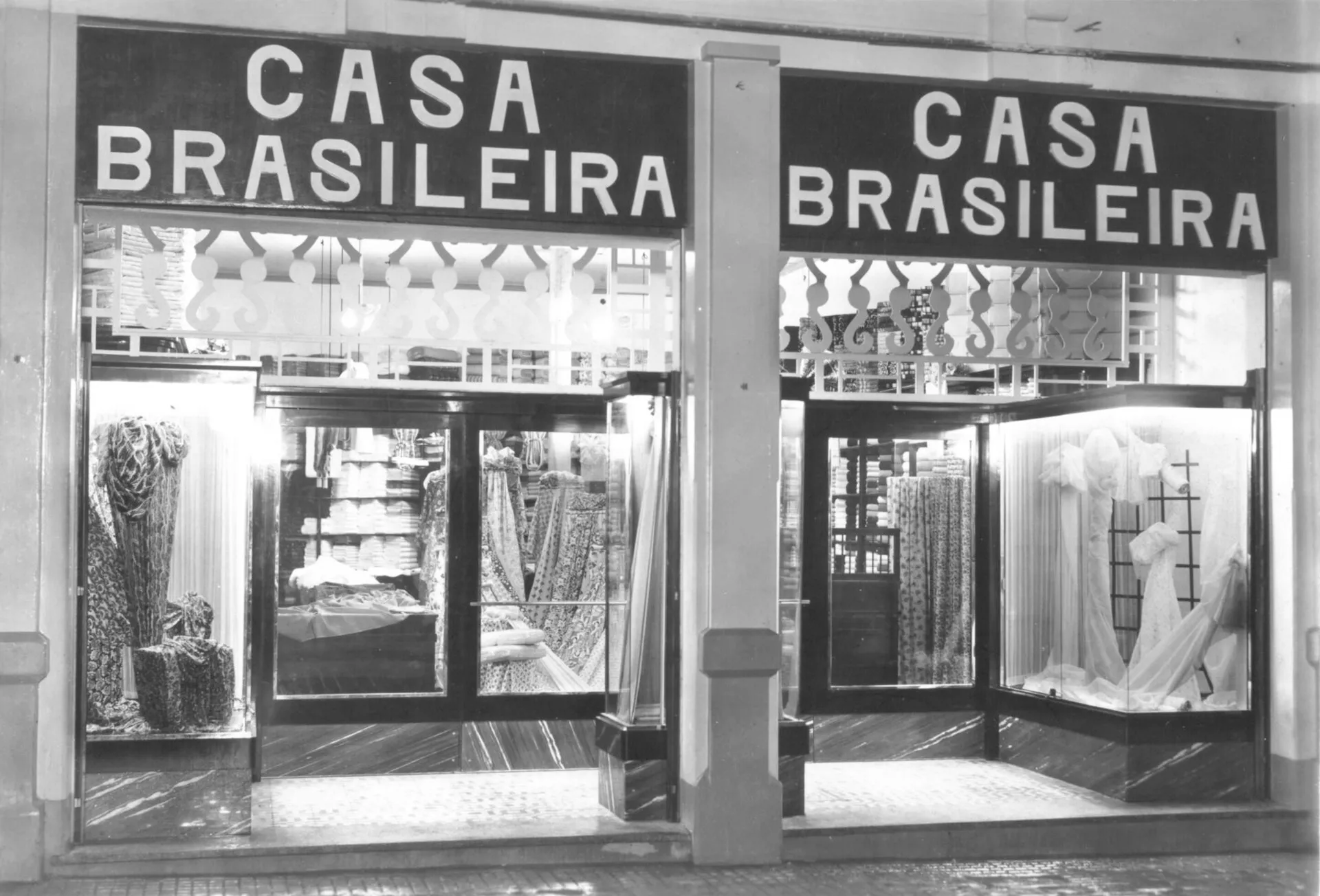 Fachada da loja Casa Brasileira, com letreiros sobre duas vitrines e tecidos expostos, em foto antiga em preto e branco.