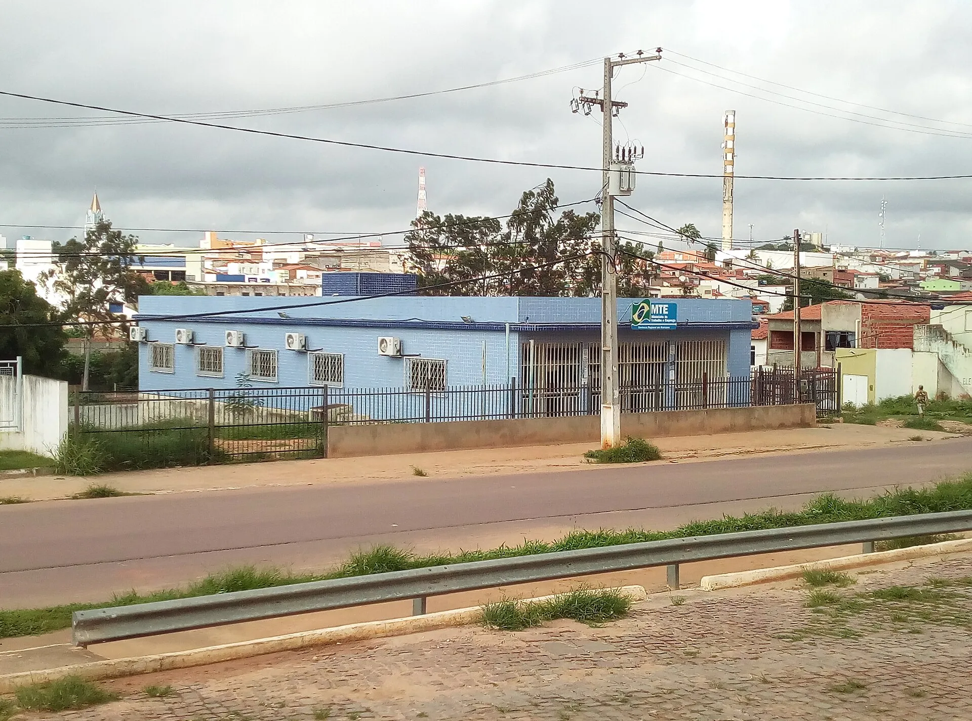 Prédio azul de um escritório do Ministério do Trabalho e Emprego, com placa verde e amarela do MTE na fachada, em rua de cidade do interior sob céu nublado.