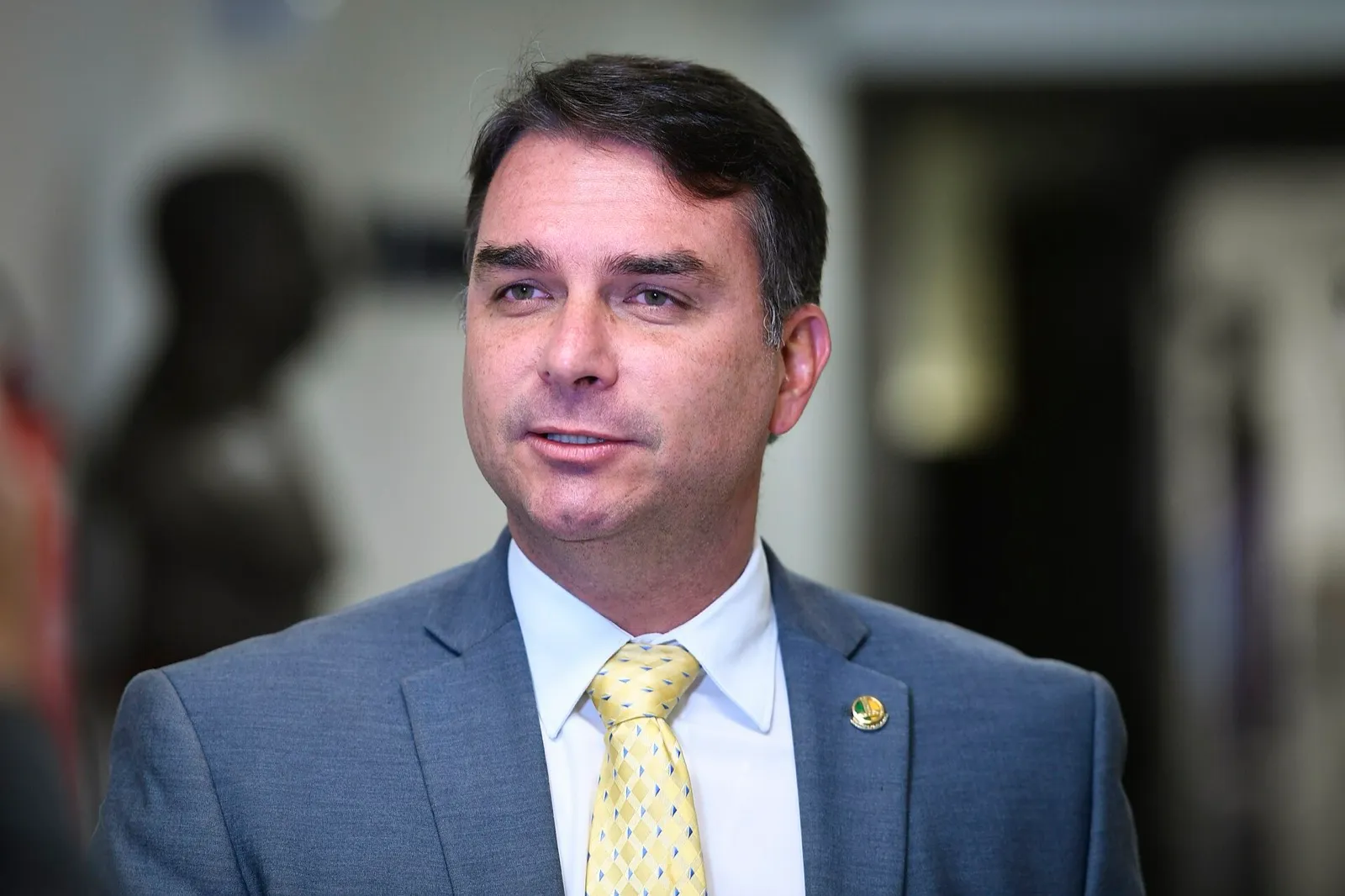 O senador Flávio Bolsonaro, de terno cinza e gravata amarela, concede entrevista em ambiente interno.