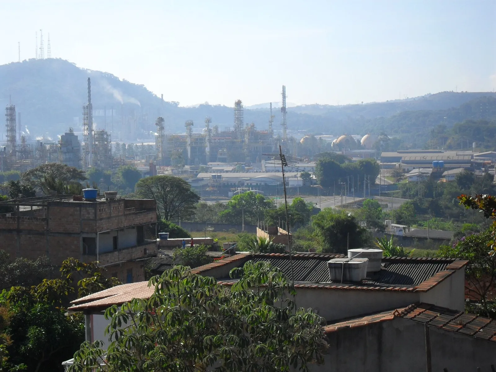 Vista de longe da Refinaria Gabriel Passos, da Petrobras, com torres, tubulações e esferas de armazenamento, atrás de casas e árvores em primeiro plano.