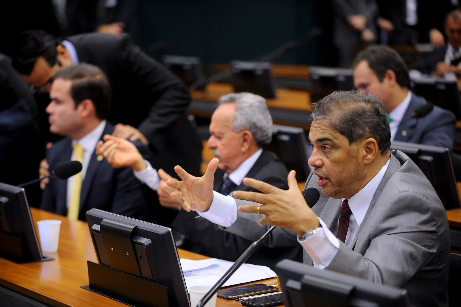 Parlamentar de terno cinza gesticula ao microfone em reunião da Comissão Mista de Orçamento, com outros parlamentares sentados ao lado.
