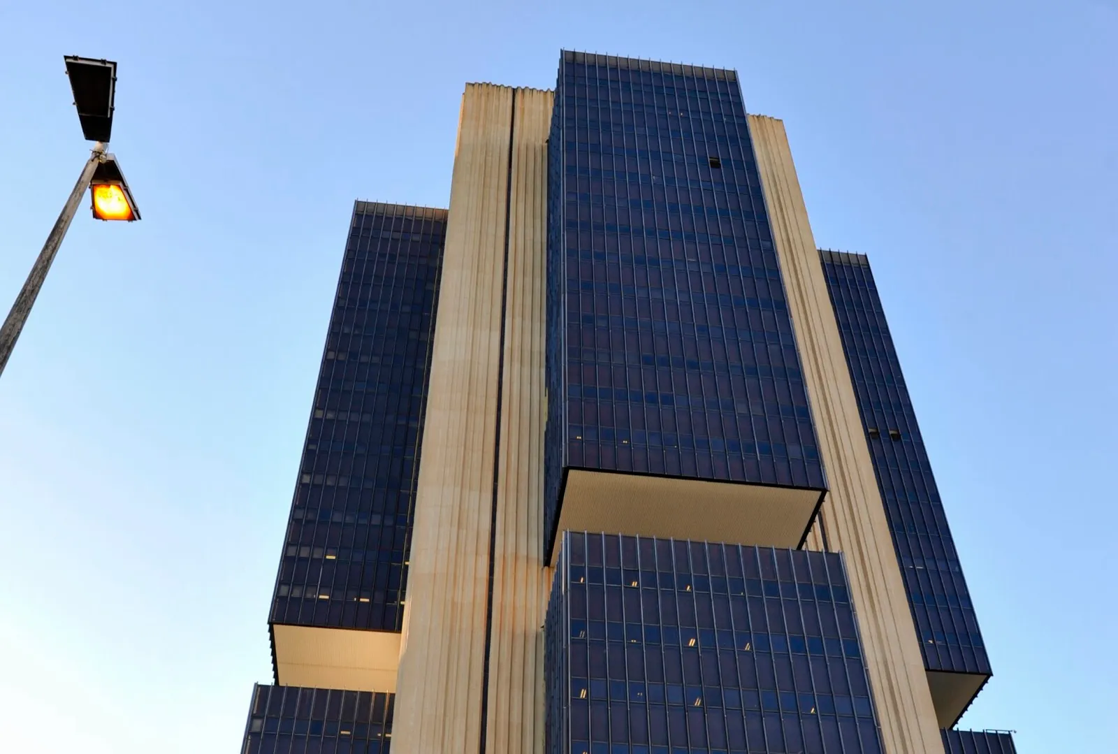 Torre da sede do Banco Central do Brasil em Brasília, com fachada de vidro azul escuro e pilares claros, vista de baixo contra o céu azul.