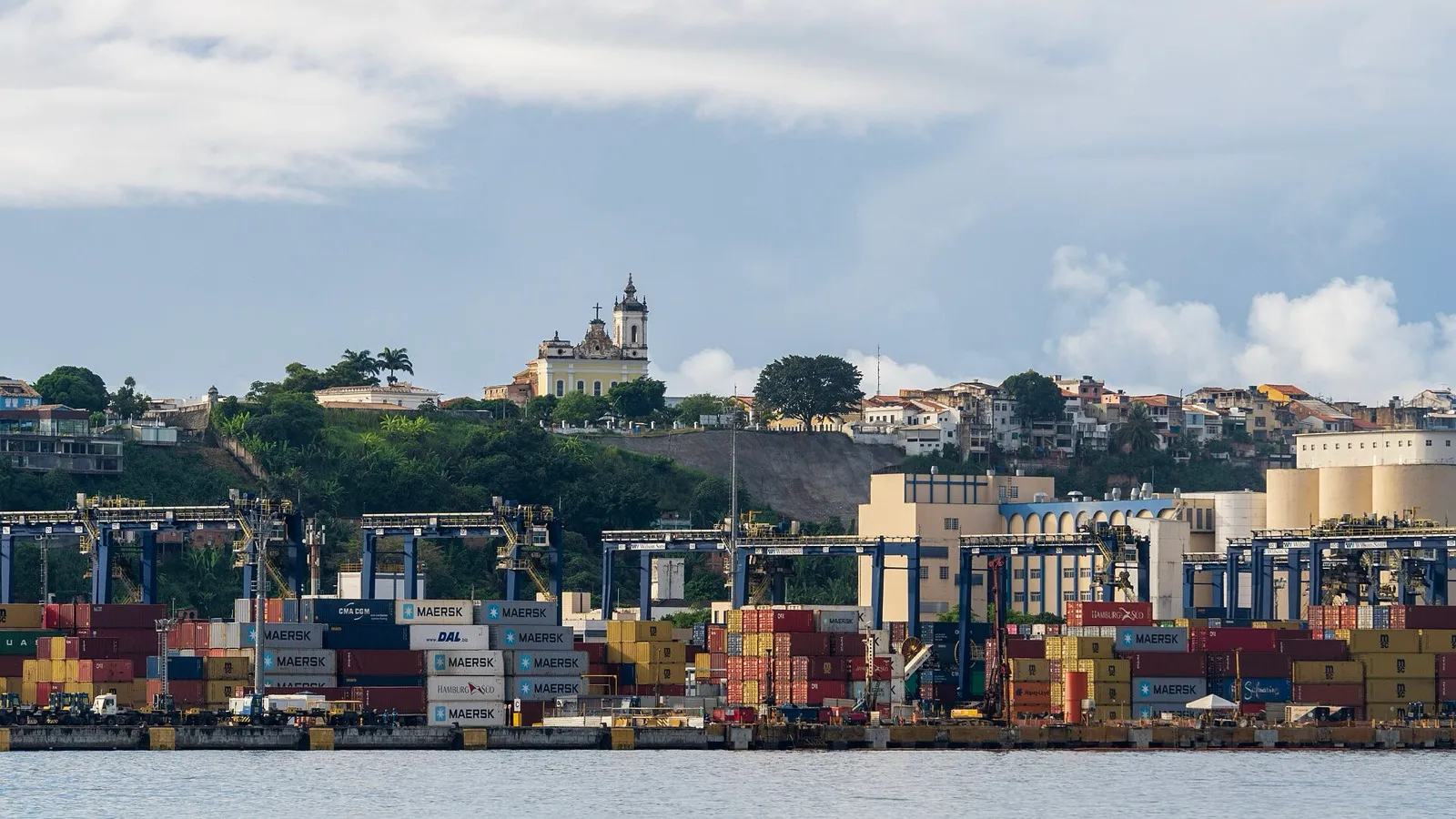 Terminal de containers em porto brasileiro, com pilhas de contêineres coloridos, guindastes azuis e uma igreja sobre o morro ao fundo.