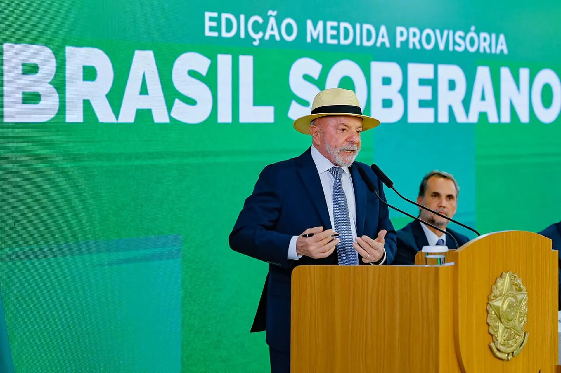 O presidente Luiz Inácio Lula da Silva, de chapéu, discursa atrás de um púlpito de madeira, diante de painel com a inscrição 'Edição Medida Provisória Brasil Soberano'